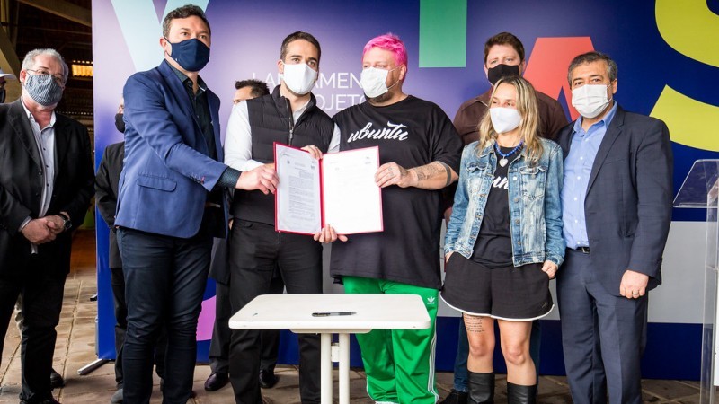Seis homens, vestidos de terno escuro e camisa clara, um deles está com camisa branca e um colete. temos na foto um rapaz de calça de abrigo verde e camiseta preta e uma menina de jaqueta jeans e saia preta. Três homens estão segurando uma ata.