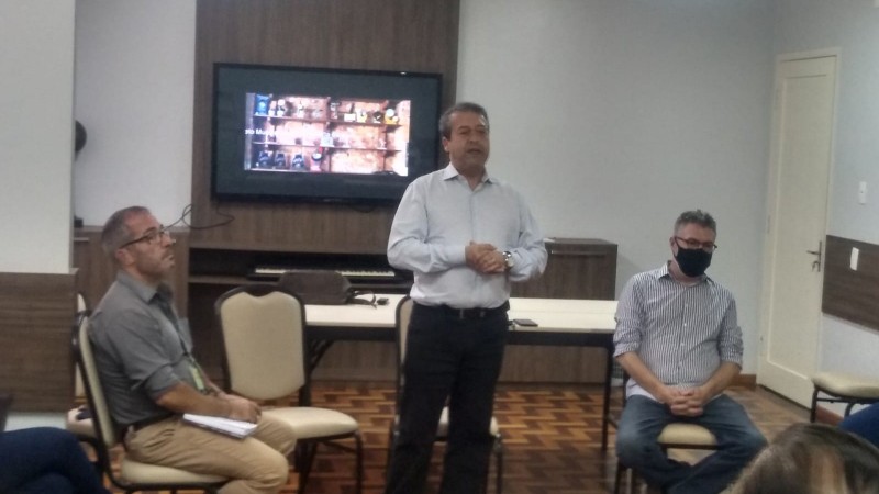 Três homens na foto, vestindo calça e camisa, dois sentados e um empé, falando.