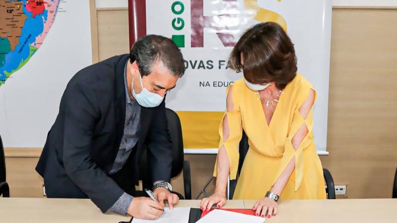 o Secretário vestido de terno escuro e a Secretária da Educação Raquel, de vestido amarelo, assinam o termo de cooperação entre secretarias. Atrás deles tem o banner do governo do Estado RS _ NOVAS FAÇANHAS