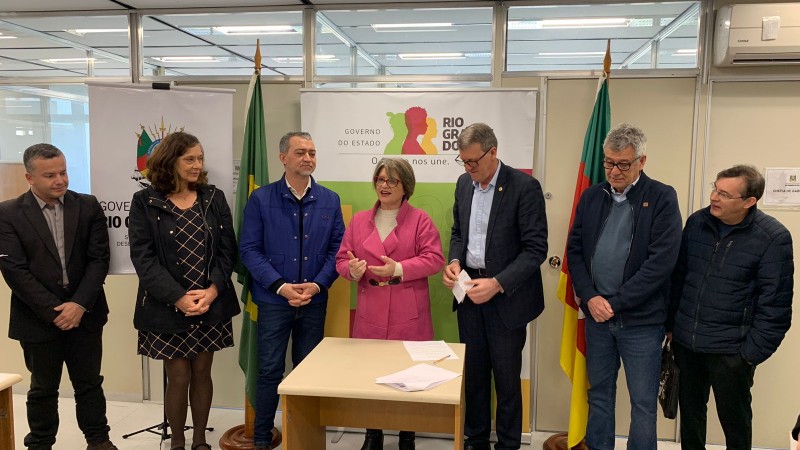 Assinatura de termo de colaboração. Pessoas enfileiradas na frente de um banner do governo do Rio Grande do Sul, uma mesa posicionada à frente com um papel e uma caneta sobre ela.