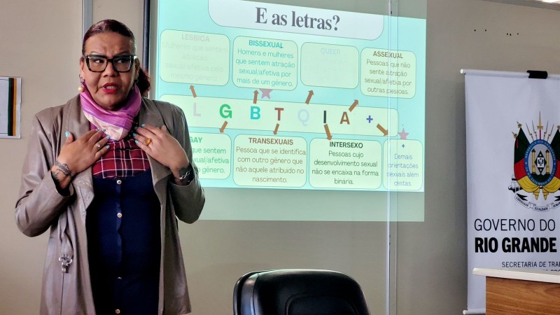 Mulher negra palestrando. Ao fundo, projeção sobre o significado das letras da sigla LGBTQIA+.