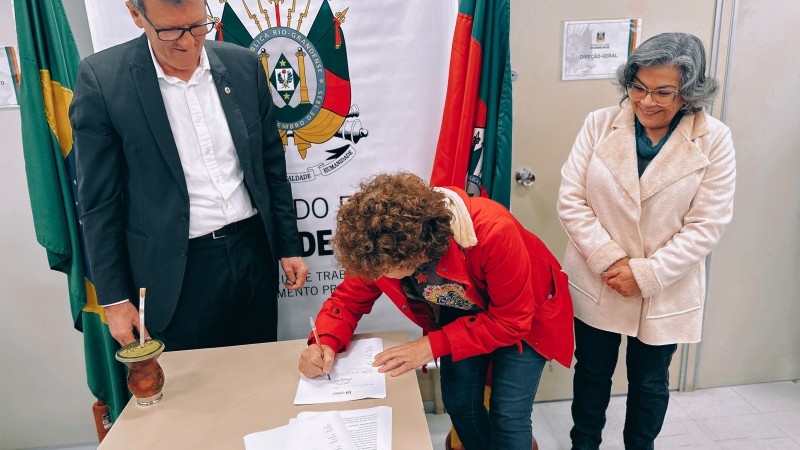Secretário Sossella, deputada Stela Farias e presidente da Unisol Nelso Nespolo estão presentes na imagem. Nelsa assina um papel que está sobre uma mesa. No fundo, há um banner com o brasão do governo estadual e as bandeiras do Brasil e do Estado, uma de cada lado.