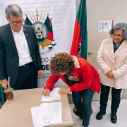 Secretário Sossella, deputada Stela Farias e presidente da Unisol Nelso Nespolo estão presentes na imagem. Nelsa assina um papel que está sobre uma mesa. No fundo, há um banner com o brasão do governo estadual e as bandeiras do Brasil e do Estado, uma de cada lado.