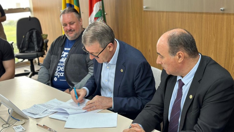 Secretário Gilmar Sossella assinando o termo de colaboração. O presidente da Avesol, Miguel Orlandi, e o deputado estadual Zé Nunes, estão sentados ao lado dele. 