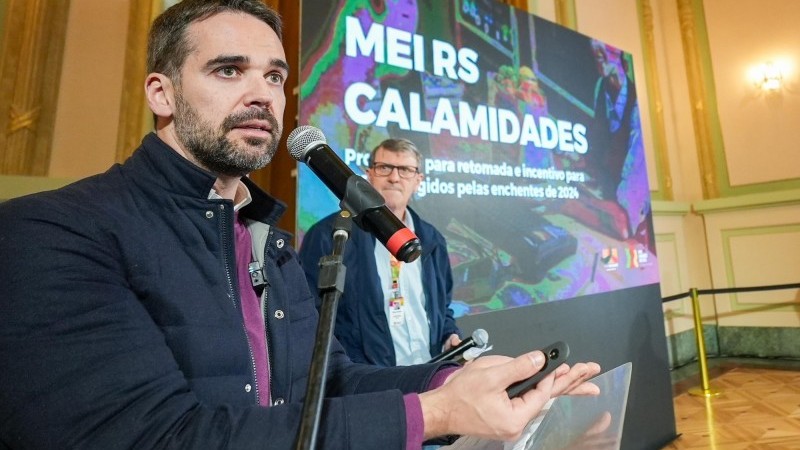 Governador Eduardo Leite está falando ao microfone. Secretário de Trabalho e Desenvolvimento Profissional está em pé, ao fundo. Há um painel escrito: MEI RS CALAMIDADES.