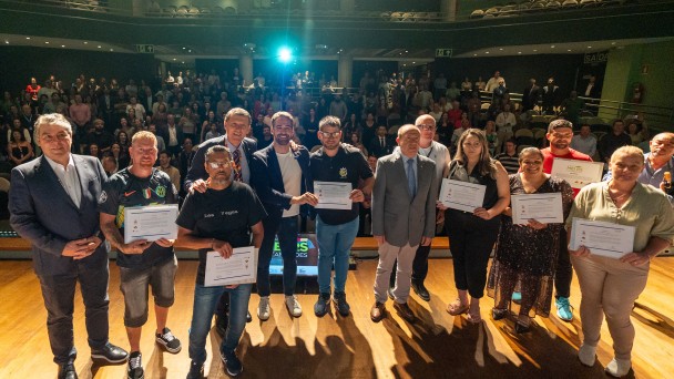 Autoridades e microempreendedores estão posando para foto no palco. Os microempreendedores seguram um certificado. Em segundo plano, uma plateia em pé. 