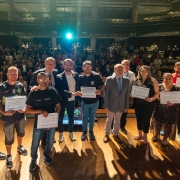 Autoridades e microempreendedores estão posando para foto no palco. Os microempreendedores seguram um certificado. Em segundo plano, uma plateia em pé. 